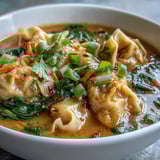 Red Curry Wonton Soup With Greens in a steamy bowl, garnished with cilantro, scallions, and bright lime wedges.