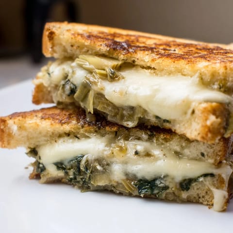 Slices of Artichoke Spinach Grilled Cheese rest on a plate, served beside a warm bowl of tomato soup.  